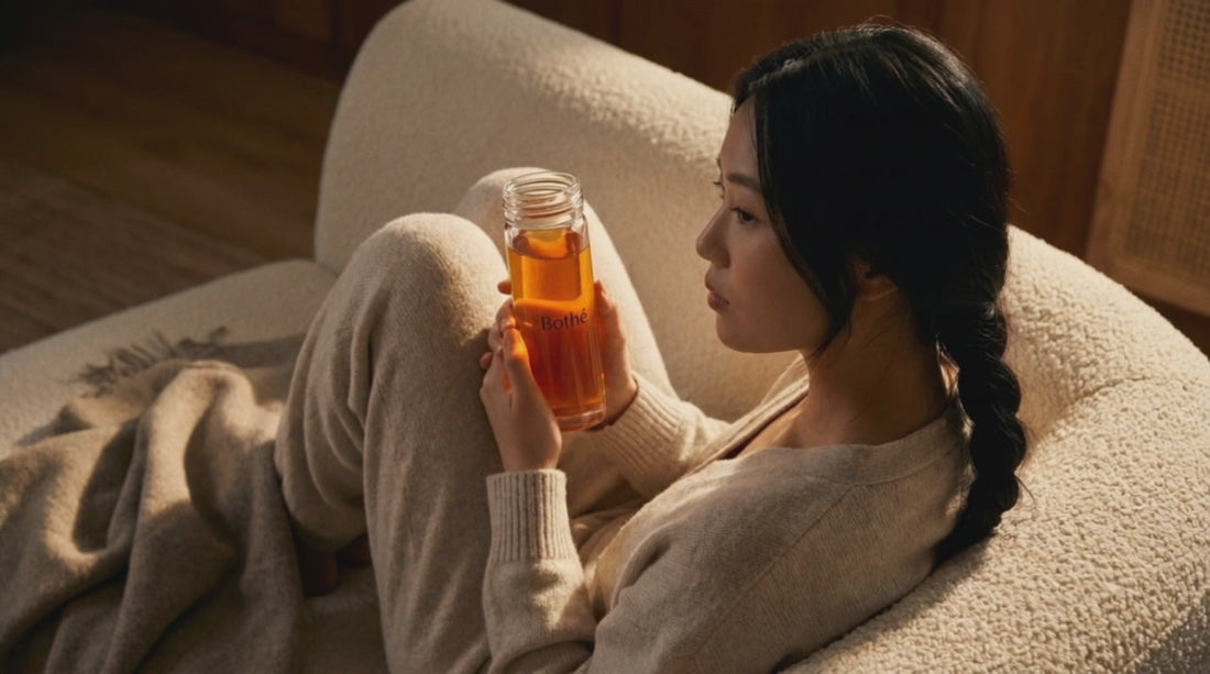 Femme lovée dans un fauteuil en fin de journée, une bouteille en verre Bothé d'infusion ambrée à la main